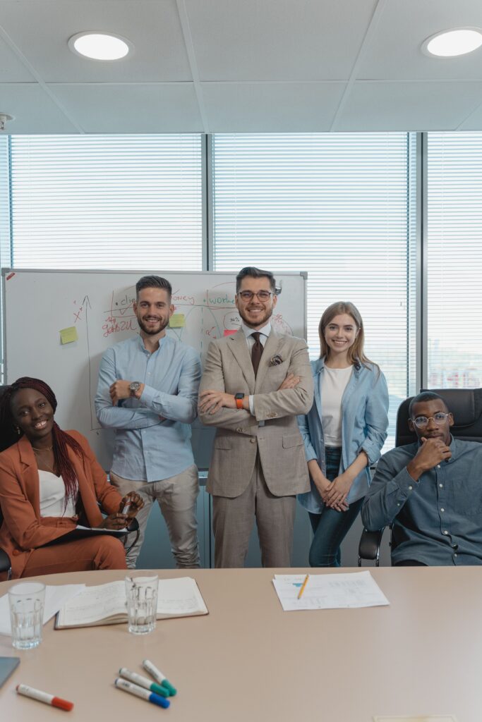 pexels-photo-5439475-5439475 Five coworkers smiling and posing in a bright modern office with a whiteboard and window view.